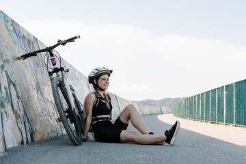 Cheerful young female in activewear and helmet sitting on asphalt path near bicycle and stretching legs while preparing for trip in summer day