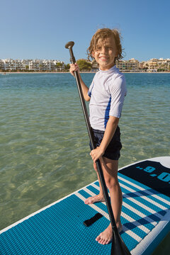 Vertical photo of the portrait of a boy standing on a paddle surfboard with a paddle facing the camera while smiling with the beach with buildings on the background