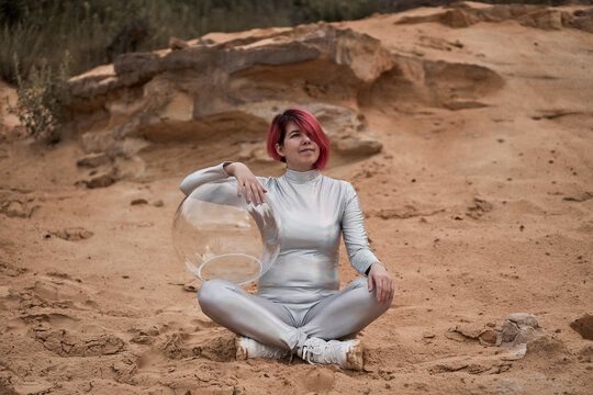 Positive young female with dyed red hair wearing silver space suit and glass helmet and looking away while sitting on rocky formation
