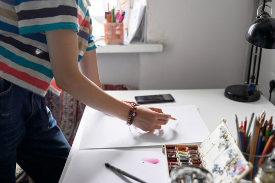 Crop anonymous female artist painting with watercolors and watching online video tutorial on smartphone while creating artwork in studio
