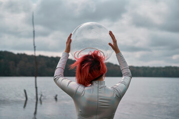 Back view of unrecognizable futuristic young red haired female in silver space suit and glass helmet walking on flooding river