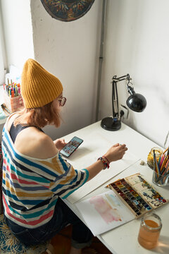 Female artist painting with watercolors and watching online video tutorial on smartphone while creating artwork in studio