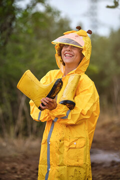 Vertical photo of a child with yellow raincoat with an animal-shaped hood holding a pair of rain boots in his hands while smiling looking ahead in the middle of a forest