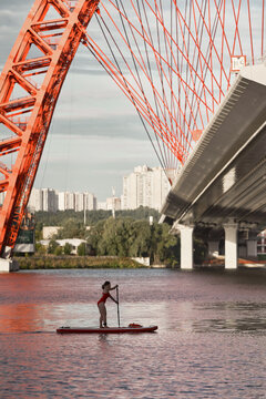Side view of woman standing on paddle board in river and rowing with paddle under bridge in city