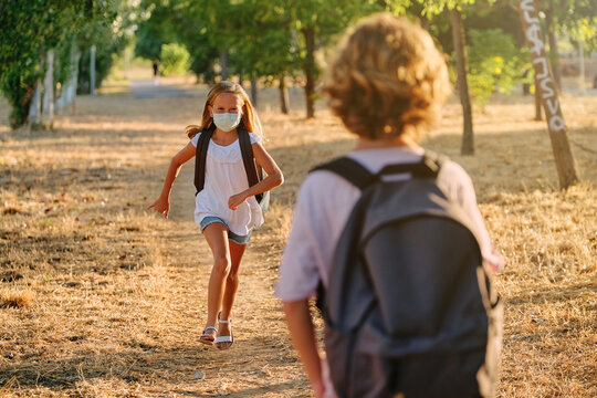 Blonde Girl With Mask And School Bag Running Towards A Boy Who Is Waiting For Her On A Path Surrounded By Trees