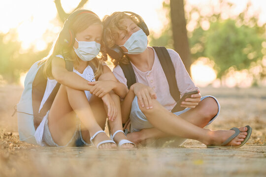 Two children with school bags and masks sitting in the woods listening to music with their heads together and emotional expression with wireless headphones connected to their mobile phones