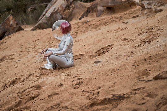 Positive young female with dyed red hair wearing silver space suit and glass helmet and looking away while sitting on rocky formation