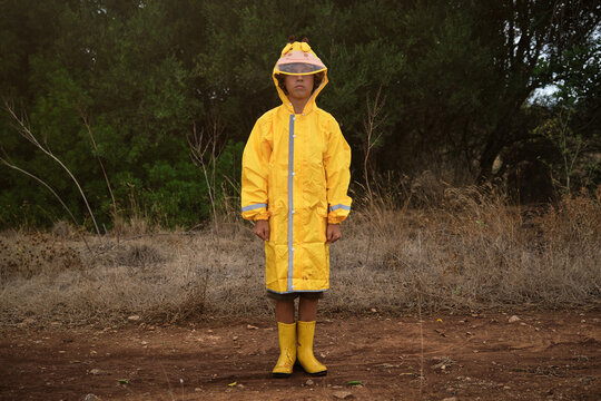 Portrait of a blond boy in a yellow hooded raincoat and yellow rain boots standing in the middle of a forest on a rainy day