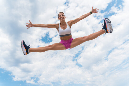 From below of strong female athlete in moment of jumping with splits and outstretched arms during workout on background of cloudy sky