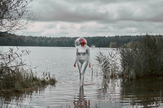 Futuristic young red haired female in silver space suit and glass helmet walking out of flooding river