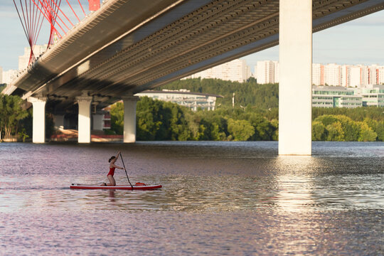 Side view of woman standing on paddle board in river and rowing with paddle under bridge in city