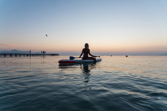 Back view of of unrecognizable female surfer sitting on paddleboard and rowing against spectacular sun in sunset sky