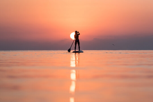 Back view of silhouette of unrecognizable female surfer standing on paddleboard and rowing against spectacular sun in sunset sky