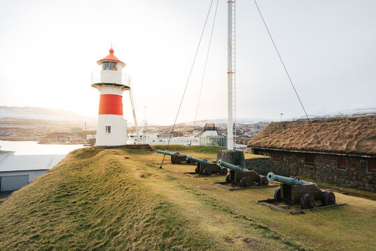 Picturesque view of shabby cannons placed in row on grassy hill with lighthouse on background of gray sky on Faroe Islands in autumn