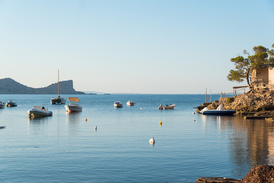Picturesque scenery of old cove near sea with moored boats on rocky shore and trees under blue sky in Sa Caleta