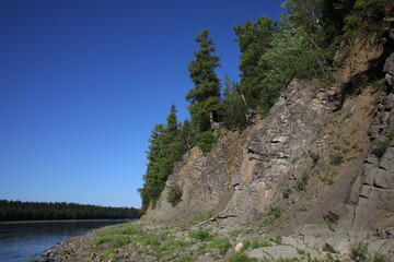 Trees grow on a steep cliff on bare rocks near the river.The blue sky is reflected on the surface of the calm water with the forest on the other side.Summer trips to the Urals.Nature background.Russia