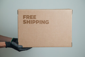 Detail of man with black gloves holding a cardboard box written free shipping to deliver products on gray background. Delivery concept. Delivery service concept. Copy space. Black friday concept.