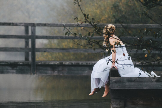 barefoot bride in beautiful white wedding dress sitting on a jetty at a lake