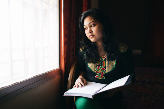 Beautiful teenage girl looking out the window with a open notebook in hand