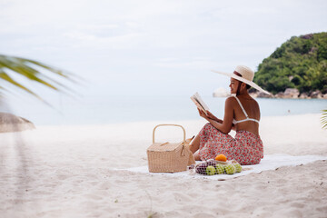Romantic portrait of  stylish asian woman sit on beach carpet blanket having picnic with basket of fruits, wearing top, long skirts and big large straw hat, holding reading book.  