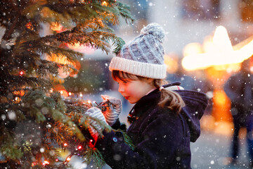 Little cute kid girl having fun on traditional Christmas market during strong snowfall. Happy child...
