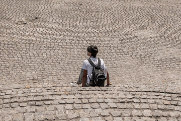 Aerial view to male tourist with a backpack rests on the stone steps against the background of a square with cobbled granite paving stones. Sitting man isolated on gray textured background