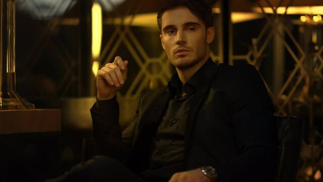 Confident man considering someone in dark room. Black suit boss sitting armchair