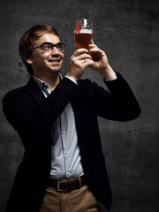 Smiling man in stylish official jacket and shirt holds up a glass of drink craft beer with both hands and looks against light