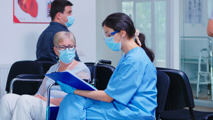 Nurse explaining diagnosis to disabled senior woman in wheelchair. Assistant wearing face mask...