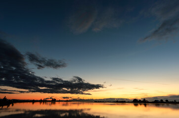 A huge sky with thick gray clouds reflecting in the river, a scarlet sunset over the black silhouettes of trees, the horn of the moon behind the clouds
