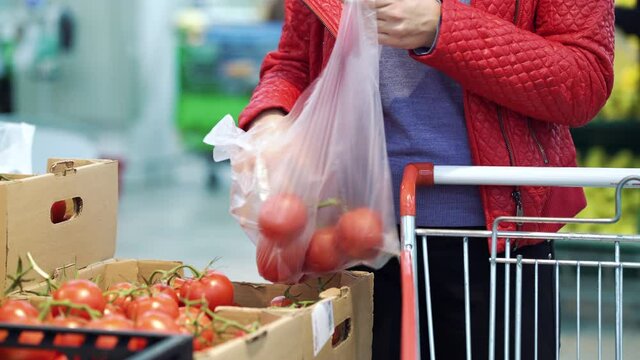 Young woman wearing red jacket picking tomatoes from box at grocery store, putting them in plastic bag and adding to trolley, blurred background. Cropped housewife buying food. Concept of shopping