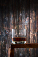 Round glass with a portion of whiskey on the counter against the old wooden wall.