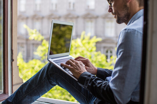 Man working on a laptop from home, sitting on a windowsill