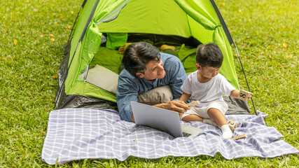 A gay is having a good time with his adopted son in front of tent checking social media with computer laptop