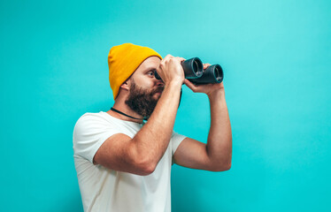 Young bearded hipster guy with binoculars over isolated blue wall wearing white blank t-shirt....