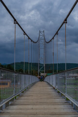 suspended pedestrian bridge with metal vertical cables and wooden path against the backdrop of village of Shamanka among Siberian mountains, cloudy sky landscape