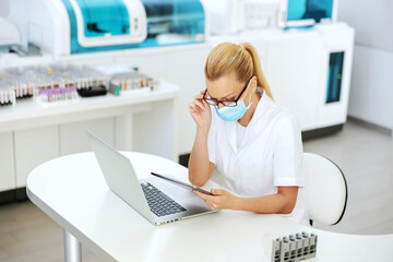 Obraz na płótnie Canvas Attractive positive blond female lab assistant in sterile uniform with surgical mask on standing in laboratory with arms crossed and looking at camera.