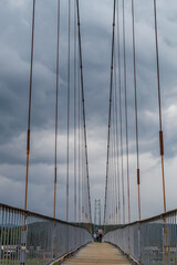 Obraz premium suspended pedestrian bridge with metal vertical ropes, cables and wooden path against the backdrop of cloudy sky, geometric, symmetrical view