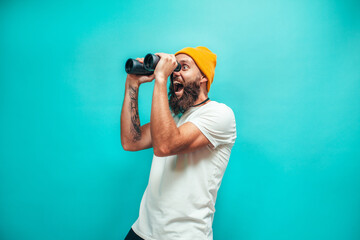 Young bearded hipster guy with binoculars over isolated blue wall wearing white blank t-shirt....