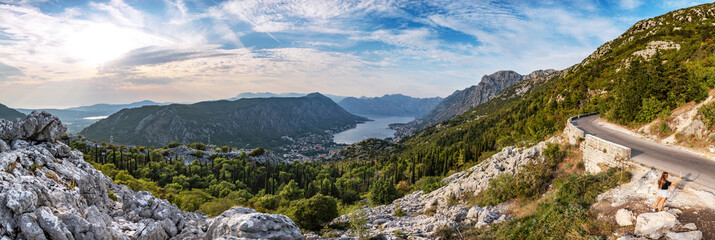 Lovely panorama of Montenegro, city of Kotor and bay from mountain serpentine road. Beautiful mountains of Montenegro above the sky with clouds at sunset. Beautiful young woman making a selfie.