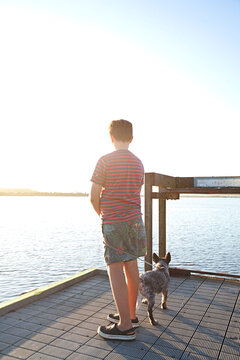A young boy standing on a pier during sunset with his puppy