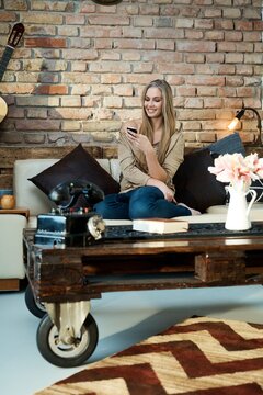 Happy blonde woman resting at home on sofa, using mobilephone, smiling.