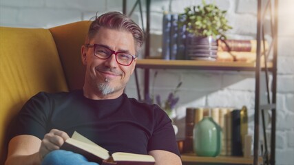 Older white man in glasses reading book at home sitting in armchair in cosy room, smiling. 