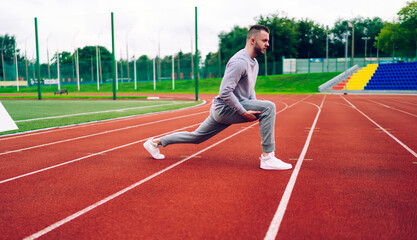 Side view of caucasian male runner stretching lower body muscles for getting better results and training flexible body, handsome young sportsman in activewear having morning workout on stadium