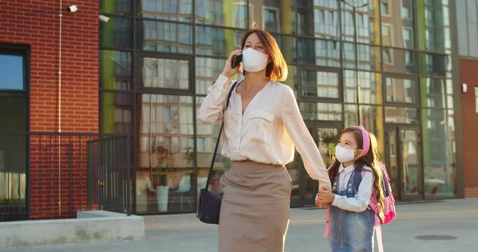 Caucasian busy woman in mask talking on smartphone while walking with cute daughter from school on sunny day. Little girl trying to bring mother's attention who is calling on cellphone School concept