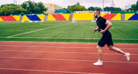 Fototapeta premium Young handsome male in earphones running on stadium dressed in active wear reaching sport goals, strong man athlete keeping fit having morning cardio training with favorite music in headphones