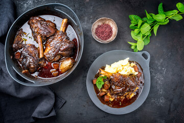 Modern style traditional braised slow cooked lamb shank in red wine sauce with shallots and mashed potatoes offered as top view on a design cast iron plate and pot