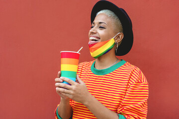 Happy young gay woman drinking from a rainbow glass while wearing a pride mask during coronavirus outbreak - Focus on girl face - obrazy, fototapety, plakaty