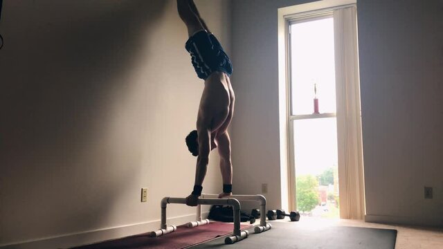 Strong young man practicing handstands on parallettes against the wall inside a sunny room