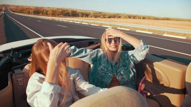 Girls in back seats of convertible take selfies on vacation. Two young women sit in car without roof shooting videos and taking photos on phone, side view